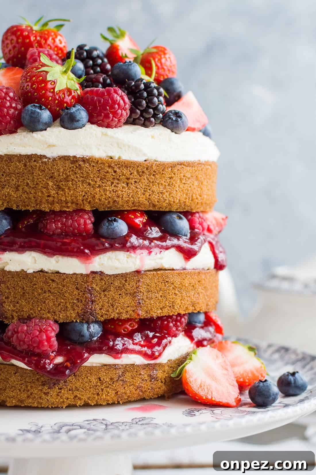 A side shot of vegan vanilla cake filled with vanilla buttercream, raspberry jam and berries on a grey background.