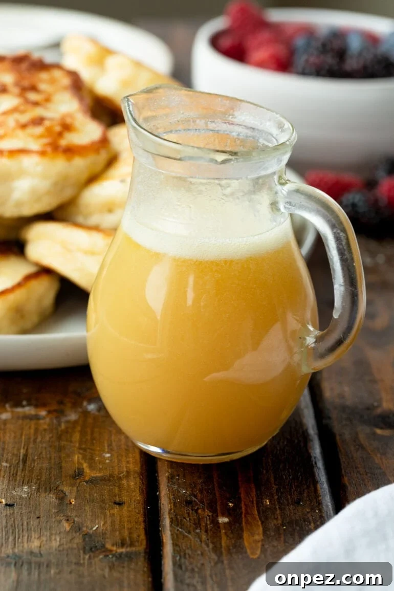 Classic Buttermilk Syrup Recipe for Pancakes and Waffles 2 a photo of a small glass pitcher full of golden buttermilk syrup with a pile of pancakes in the background