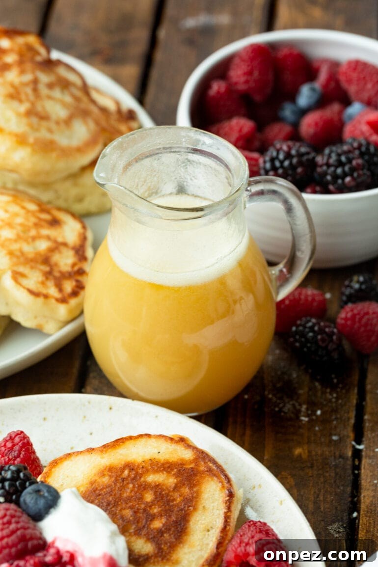 Classic Buttermilk Syrup Recipe for Pancakes and Waffles 6 a photo of a small glass pitcher of golden buttermilk syrup with a bowl of fresh berries and a plate of pancakes in the background.