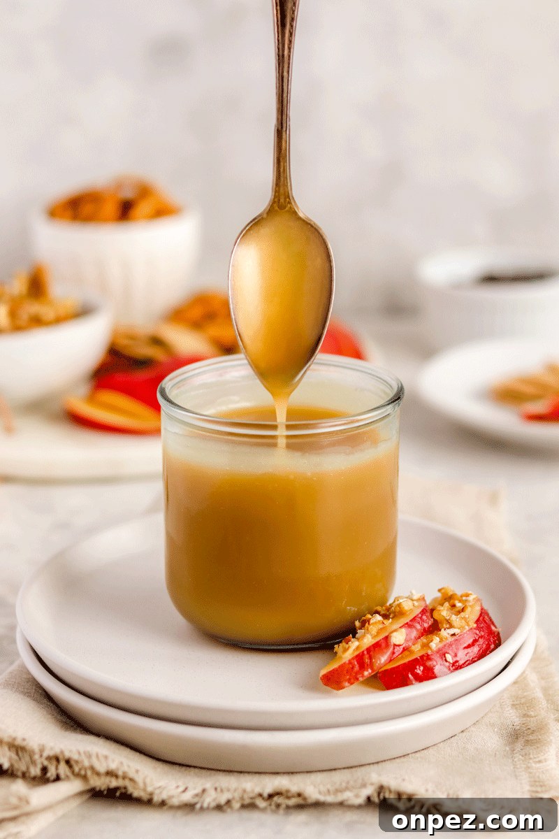 caramel sauce in a jar on a plate with apple slices on it. There is a spoon drizzling the caramel into the jar.