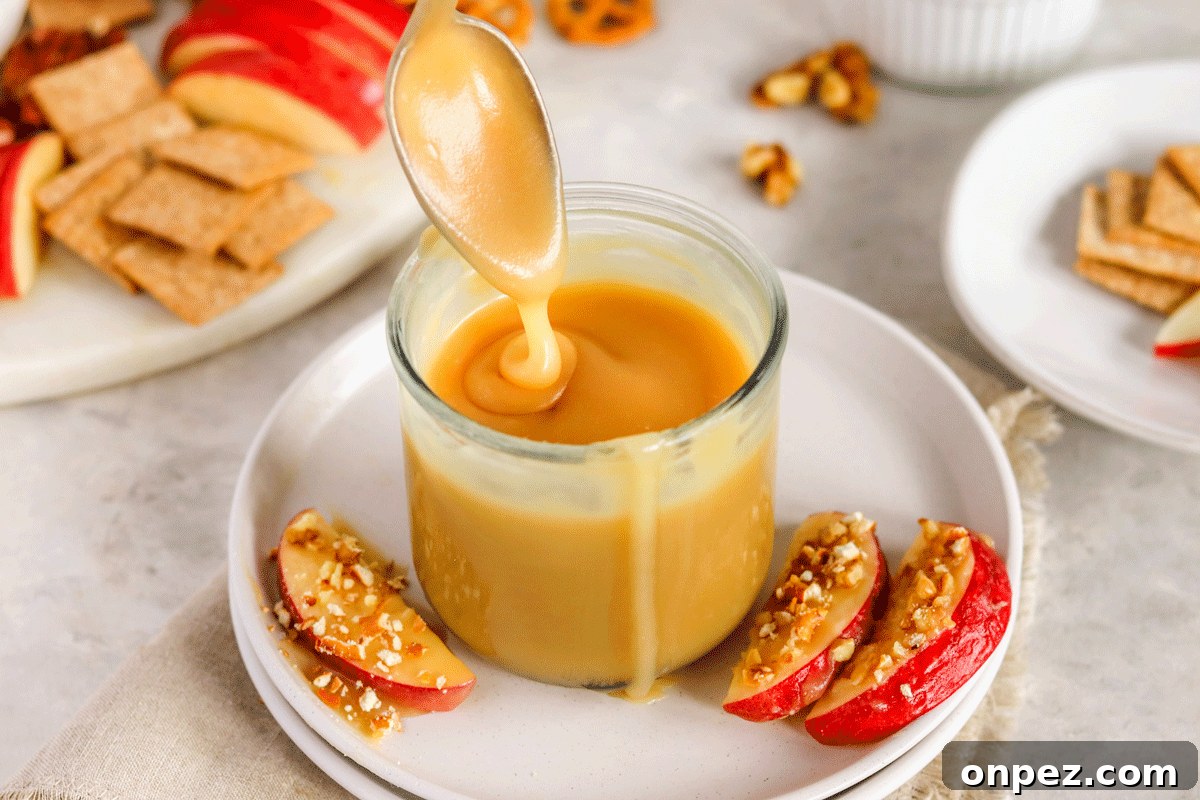 caramel sauce in a jar on a plate with apple slices on it. There is a spoon drizzling the caramel into the jar.