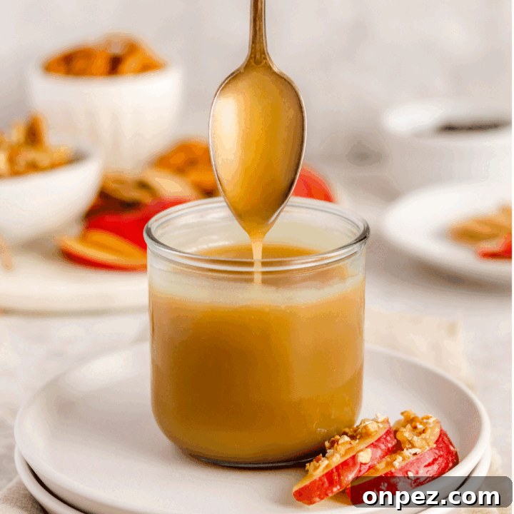 caramel sauce in a jar on a plate with apple slices on it. There is a spoon drizzling the caramel into the jar.