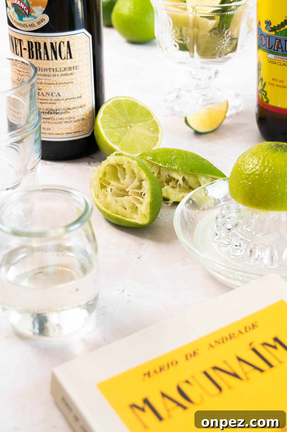juiced limes with a glass citrus juicer, some liquor bottles and the edge of the Macunaíma book on a white surface