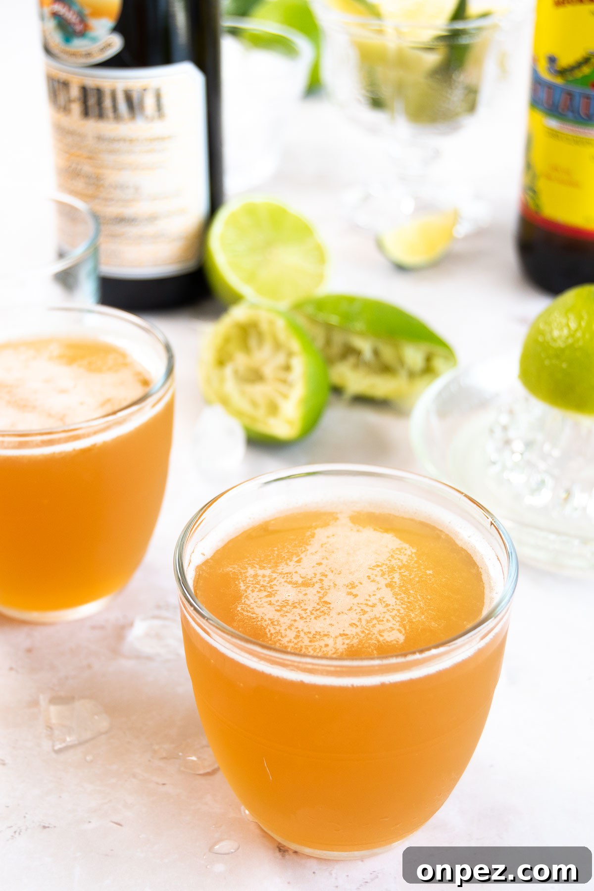 side angle of two glasses of a Macunaíma drink in small glasses near bottles of liquor and juiced limes on a white surface
