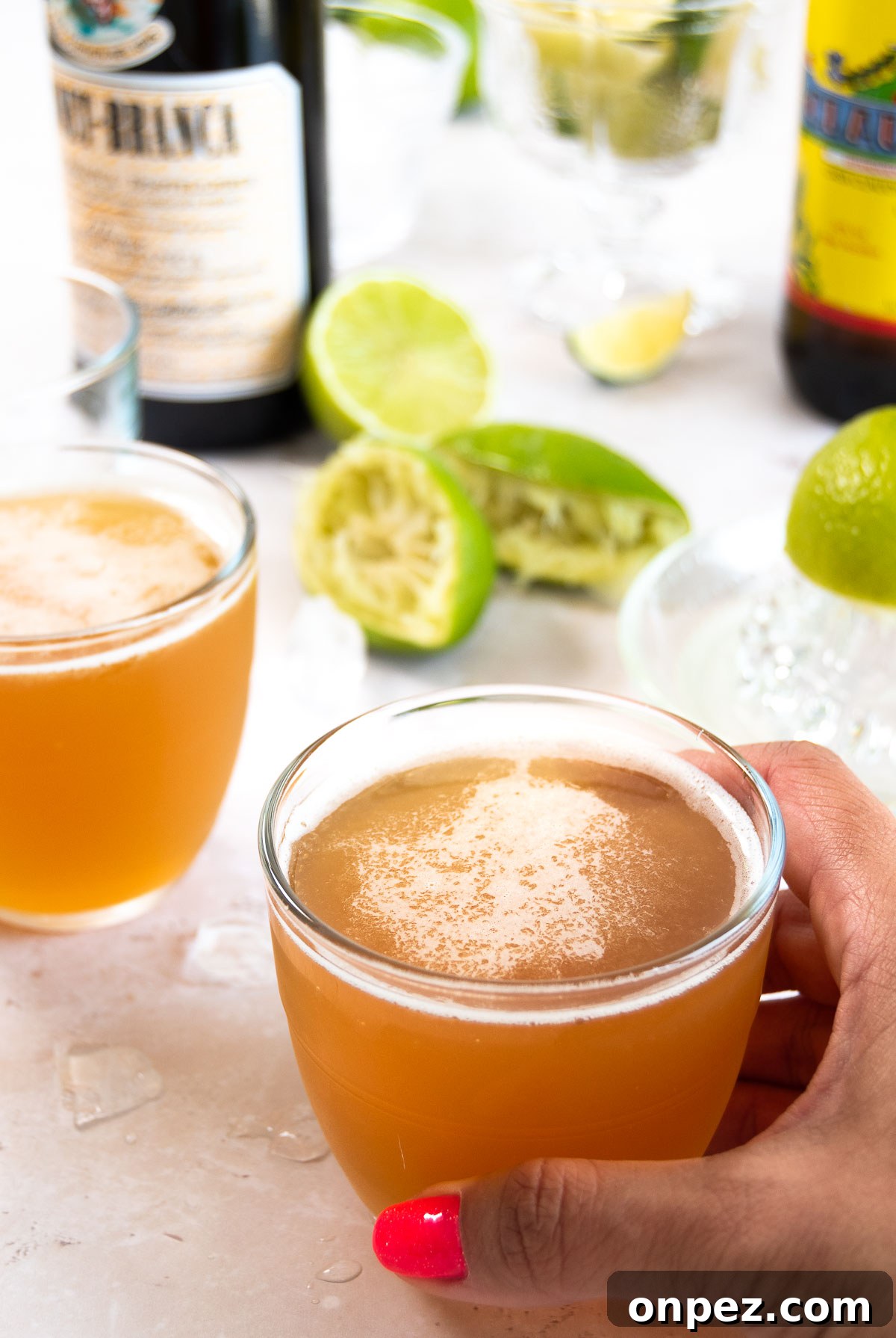 a hand holds a small glass holding the Macunaima drink next to another glass of the drink, juiced limes, ice and liquor bottles on a white surface