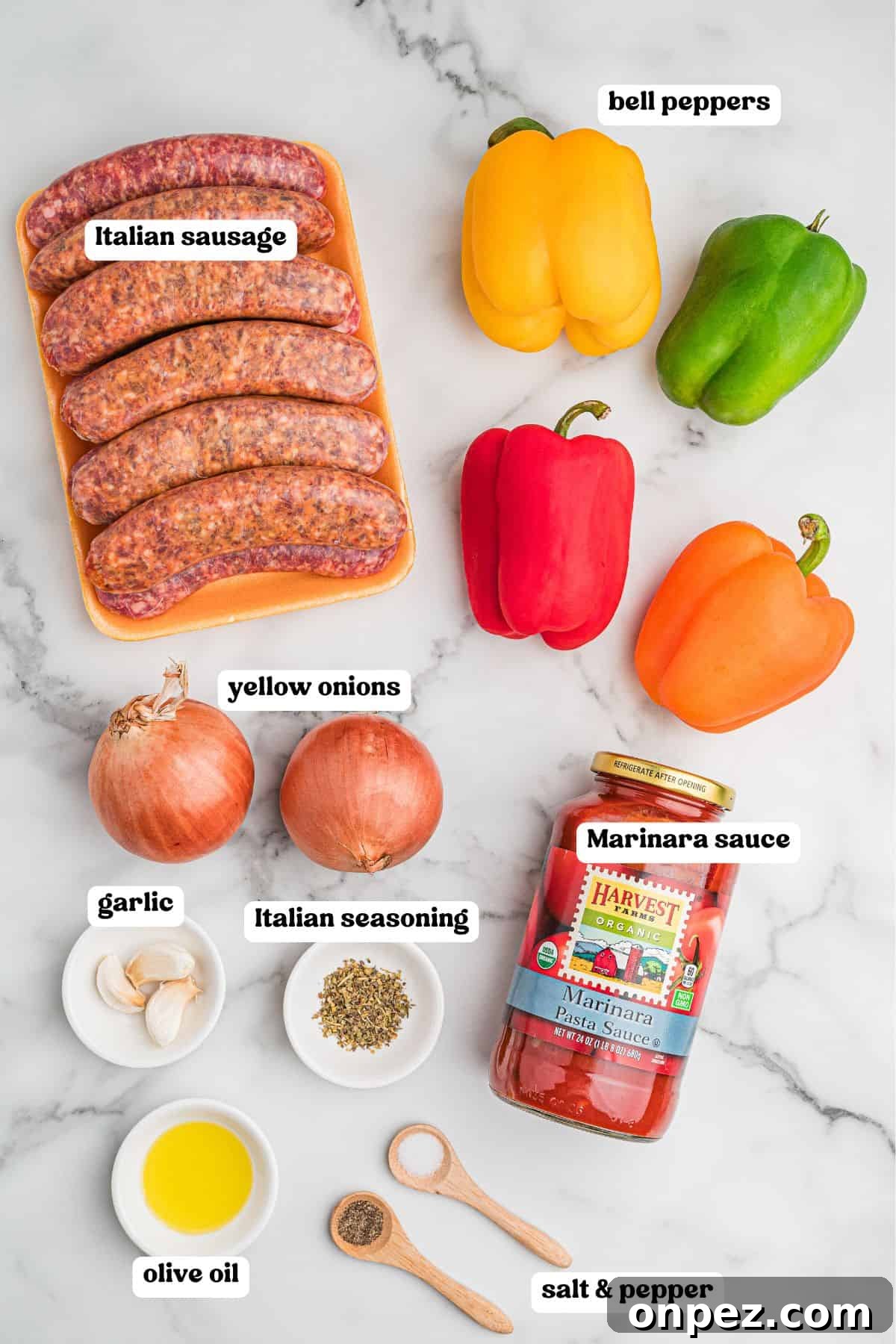 Ingredients for sausage and peppers laid out on a marble surface with labels: a pack of raw Italian sausages, yellow, green, red, and orange bell peppers, two yellow onions, garlic cloves, Italian seasoning, a jar of marinara sauce, a small dish of olive oil, and wooden spoons holding salt and pepper.