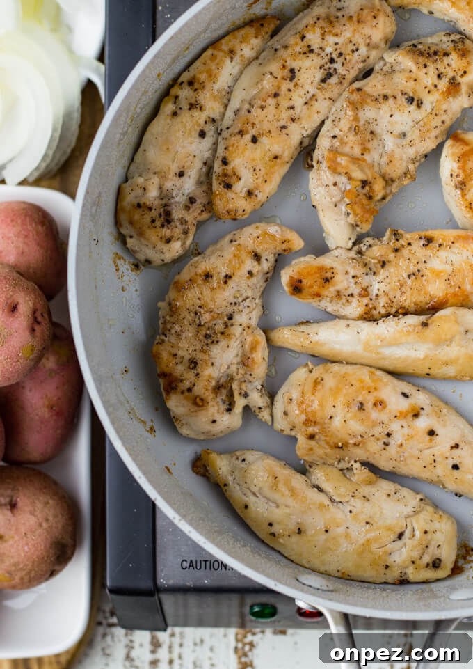 A skillet with pan fried chicken tenders with whole potatoes and sliced onions sit next to it.