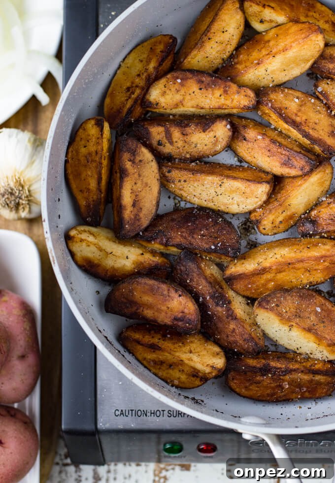 A skillet with fried potato wedges. Whole red potatoes, garlic bulbs and sliced onions sit next to the skilled.