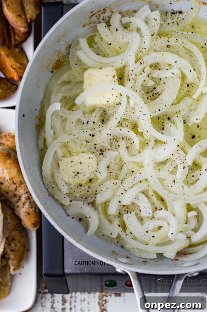 A skillet of sliced onions sit in a pan with cubes of butter melting in them. Pan fried potato wedges and chicken tenders sit next to the skillet.