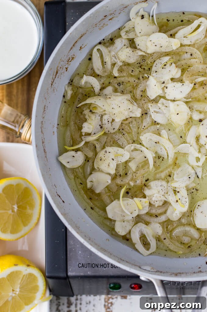A skillet of sauteed onions with sliced garlic in them sit in a wine sauce. Lemon halves and salt sit next to the skillet.