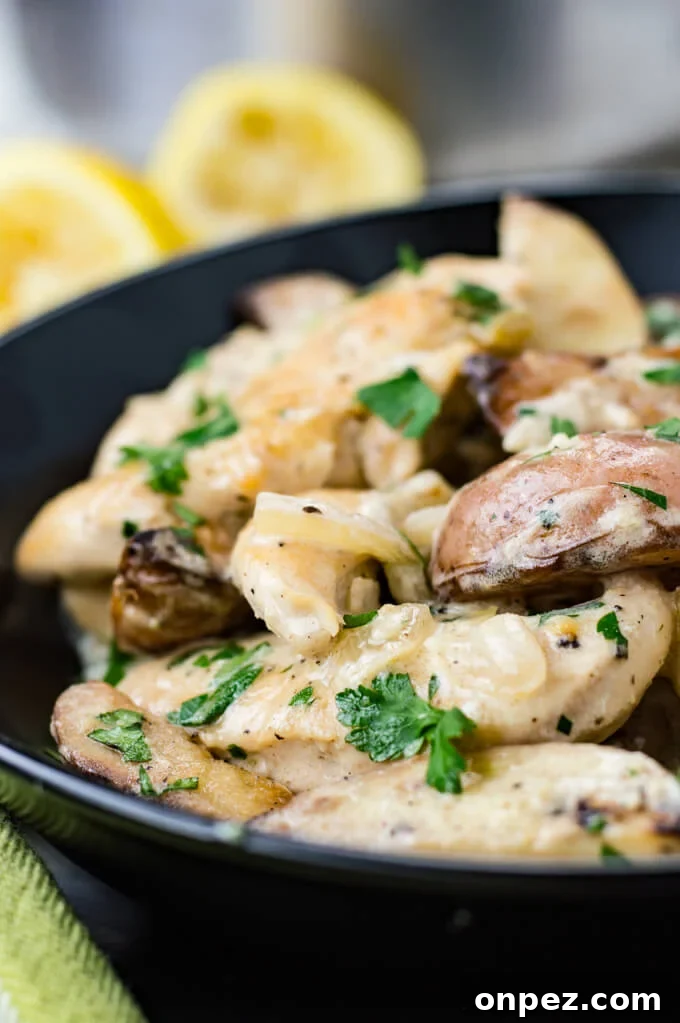 Chicken tenders and potato wedges are in a creamy lemon sauce and sitting in a black bowl with squeezed lemons in the background and a green napkin next to the bowl.
