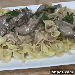 A rectangular white plate with egg noodles topped with creamy beef stroganoff, garnished with fresh parsley.